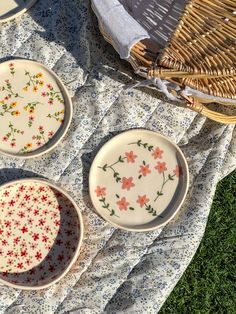 three plates with flowers on them are sitting on a blanket next to a wicker basket