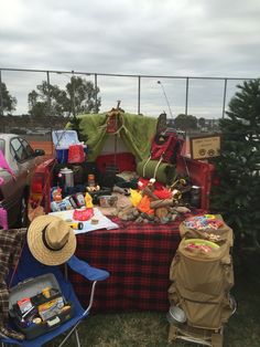 a picnic table with many items on it in the grass next to a fence and parked cars