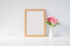 a white vase with pink flowers next to a wooden frame on a shelf against a wall