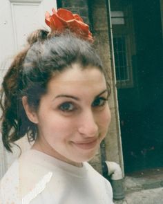 a woman with a red flower in her hair smiling at the camera while standing next to a building