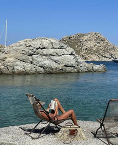 a woman sitting on top of a chair next to a body of water with rocks in the background