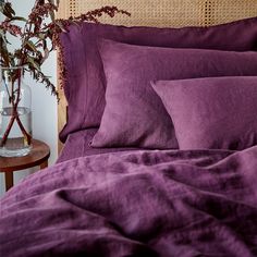 a bed topped with purple sheets and pillows next to a vase filled with flowers on top of a wooden headboard