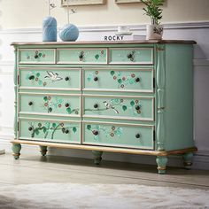 a green dresser with flowers painted on the top and bottom, sitting in a living room