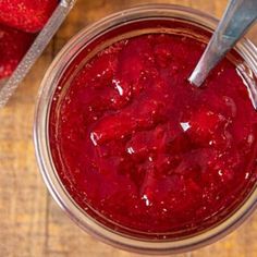 a glass jar filled with strawberry jam on top of a wooden table next to two silver spoons