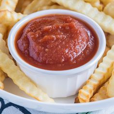 a white plate topped with waffle fries covered in ketchup next to a bowl of sauce