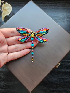 a hand holding a colorful dragon brooch on top of a metal surface next to a plant