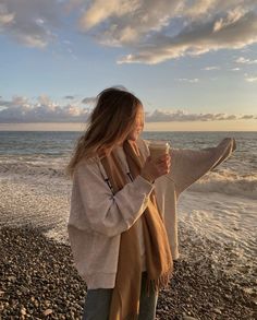 a woman standing on top of a beach next to the ocean holding a coffee cup