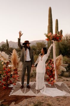 a man and woman standing in front of a cactus with their arms up while holding flowers