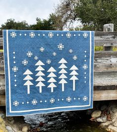 a blue quilt with white trees on it hanging from a wooden bridge over a stream