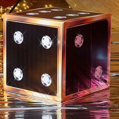 a shiny cube shaped object sitting on top of a reflective surface with lights in the background