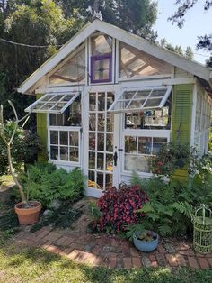 a small white house with green shutters and flowers in the front yard, surrounded by greenery