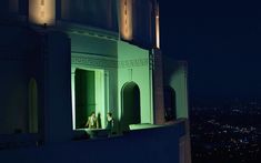 two people standing on the ledge of a building at night with city lights in the background