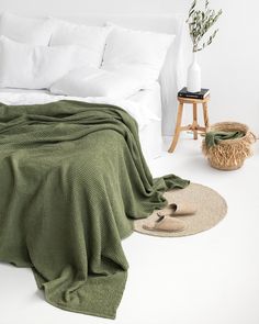 a bed with white sheets and green blanket next to a small basket on the floor