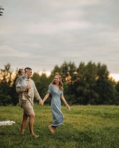 a man and woman hold hands as they walk through the grass in front of trees