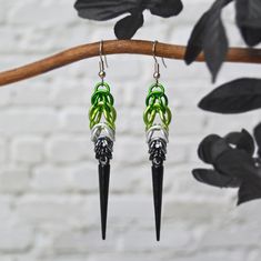 two black and green earrings hanging from a branch with leaves in front of a white brick wall