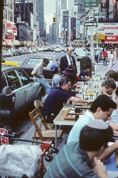 a group of people playing chess in the middle of a busy city street with tall buildings