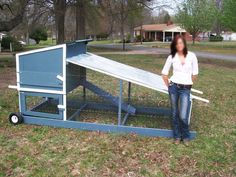 a woman standing next to a chicken coop in a yard with grass and trees behind her