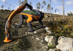 an orange and black robot standing on top of a rocky hillside next to trees, grass and rocks