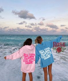 two women standing on the beach with their arms in the air while wearing matching sweatshirts