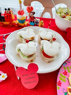 some sandwiches are sitting on a plate near a bowl of salad and mickey mouse cupcakes