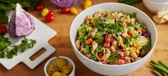 a white bowl filled with pasta salad next to other vegetables on a cutting board and utensils