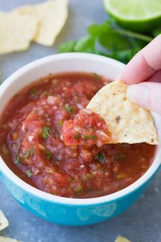 a hand holding a tortilla chip over a bowl of homemade salsa