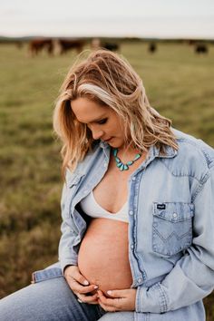 a pregnant woman sitting in a field with her stomach exposed and wearing a blue jean jacket