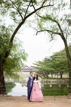 a man and woman standing next to each other in front of trees with the water behind them