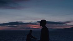 two people standing next to each other near the ocean at sunset or sunrise with mountains in the background