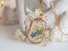 "This exquisite handmade dried flower pendant is a beautiful piece of jewelry that embodies delicacy and nature in its purest form. The pendant is made of gold-plated stainless steel, with a rectangular shape and rounded edges, giving it an elegant and sophisticated appearance. The carefully selected and arranged dried flowers, consisting of small white flowers and small blue pressed \"forget-me-not\" flowers, create a soft and elegant pattern that creates an impressive visual effect and evokes Nature-inspired Gold Flower Pendant Necklace, Gold Pressed Flower Necklace Gift For Her, Resin Flower Necklace, Pressed Flower Jewelry, Forget Me Not Flowers, Real Flower Jewelry, Transparent Resin, Fairy Necklace, Small White Flowers