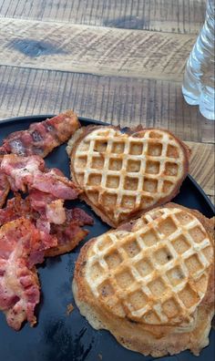 three waffles and bacon on a black plate next to a glass of water