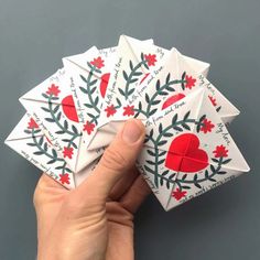 a hand holding four folded cards with red hearts and leaves on them, in front of a gray background