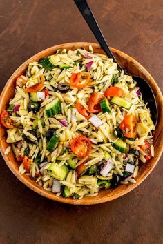 a wooden bowl filled with pasta salad on top of a brown table next to a black spoon