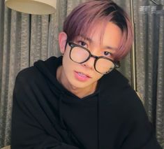 a young man with pink hair and black glasses looking at the camera while sitting in front of a window