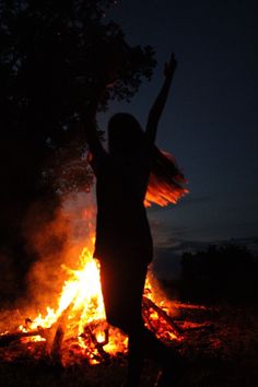 a woman standing in front of a fire with her arms up and hands raised above her head