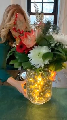 a woman arranging flowers in a vase on a table with christmas lights and garlands