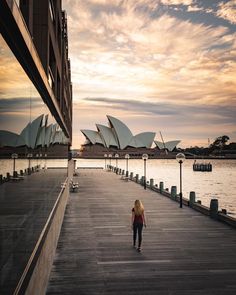 a woman walking down a wooden walkway next to the water with opera buildings in the background