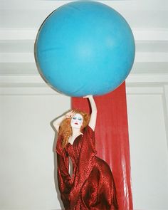 a woman in a red dress is holding a blue balloon above her head while standing next to a red curtain