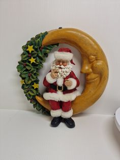 a santa clause figurine sitting on top of a crescent next to a christmas wreath