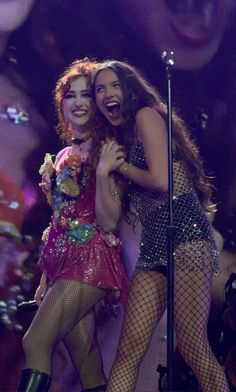 two women dressed in fishnet outfits and high heels on stage, one with her arm around the other's shoulder