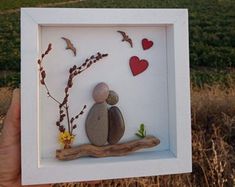 a person holding up a small frame with rocks and flowers on it in front of a field