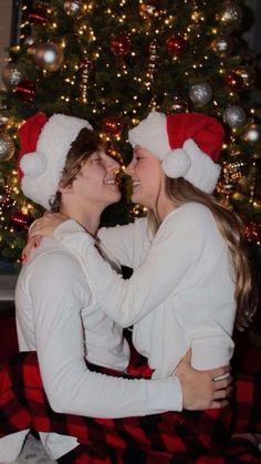 two people sitting in front of a christmas tree with their arms around each other and smiling