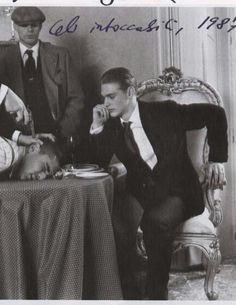 three men in suits sitting at a table talking on the phone while another man sits next to them