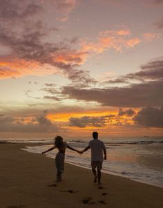 a man and woman walking on the beach holding hands as the sun sets behind them