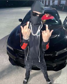 a man standing in front of a black car wearing a hoodie and holding up two fingers