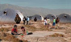 several people are standing and sitting in the desert with large metal domes behind them that look like they have been built on land