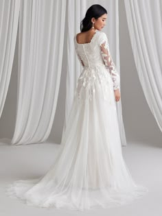 a woman in a white wedding dress with sheer sleeves and flowers on the back, standing next to curtains