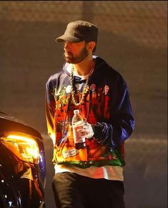 a man with a beard wearing a hat and holding a bottle in his hand while standing next to a car