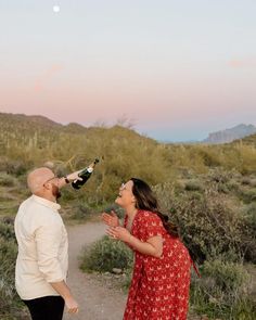 a man and woman standing on a dirt road in the desert drinking wine from bottles