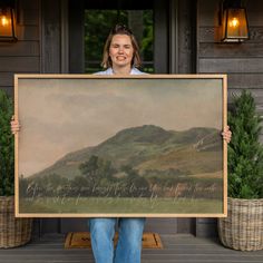 a woman holding up a large painting in front of a house with potted plants